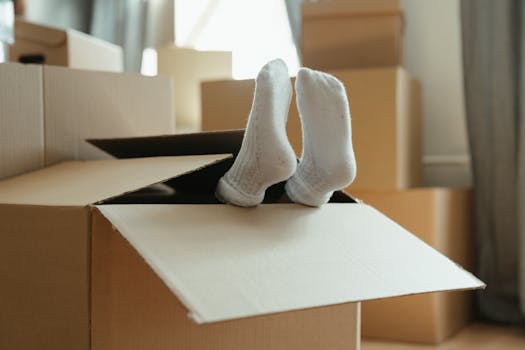 A woman lies on the floor surrounded by cardboard boxes in a dim sunlit room, expressing fatigue.
